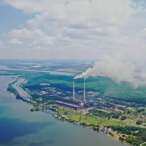 huge manufactory plant among natural background. smoke comes from pipes into the atmosphere near the river and green forest outdoors. aerial view.