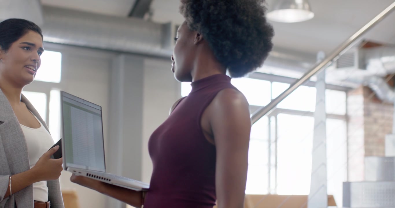 Personnel féminin diversifié vérifiant une table dans un bureau, sur le palier, montrant un ordinateur portable à une personne qui écoute au téléphone.