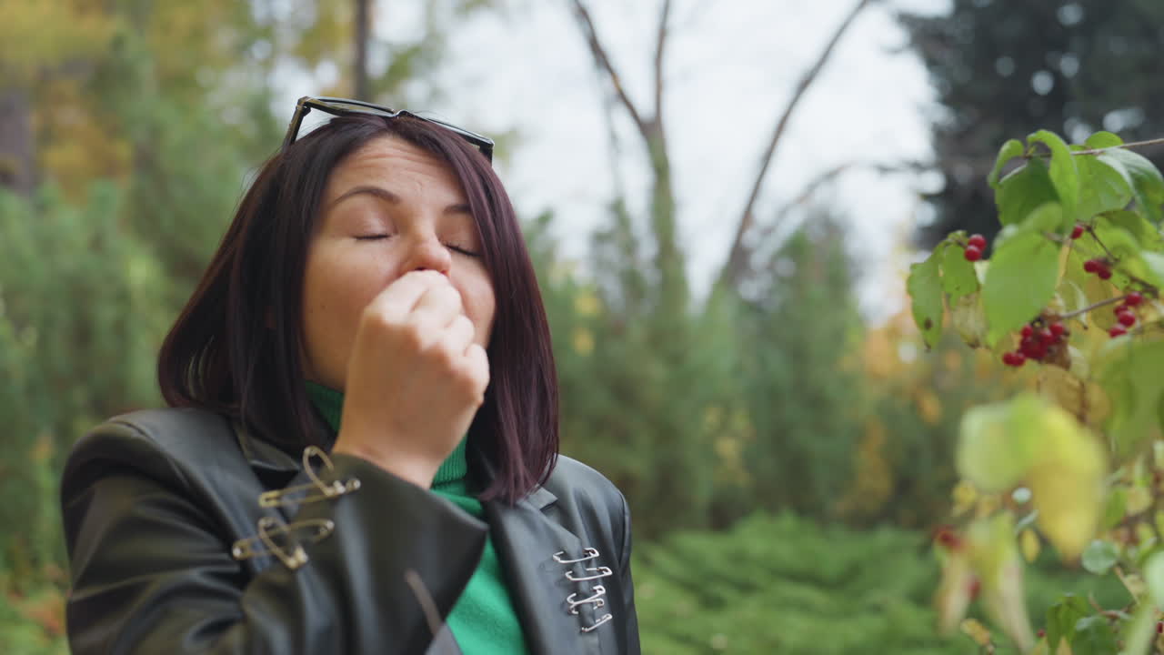 Mulher de jaqueta de couro provando frutas silvestres, close-up espontâneo em parque arborizado, óculos de sol na cabeça, folhagem de outono e arbustos verdes, expressão pensativa, moda casual com toque rocker, serena