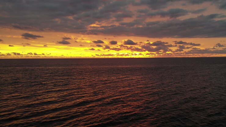 Aerial flight backwards over calm ocean with a dramatic bright yellow, orange and purple sunset