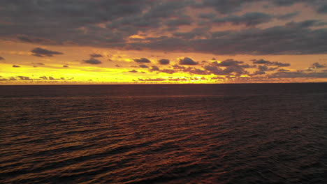 aerial flight backwards over calm ocean with a dramatic bright yellow, orange and purple sunset