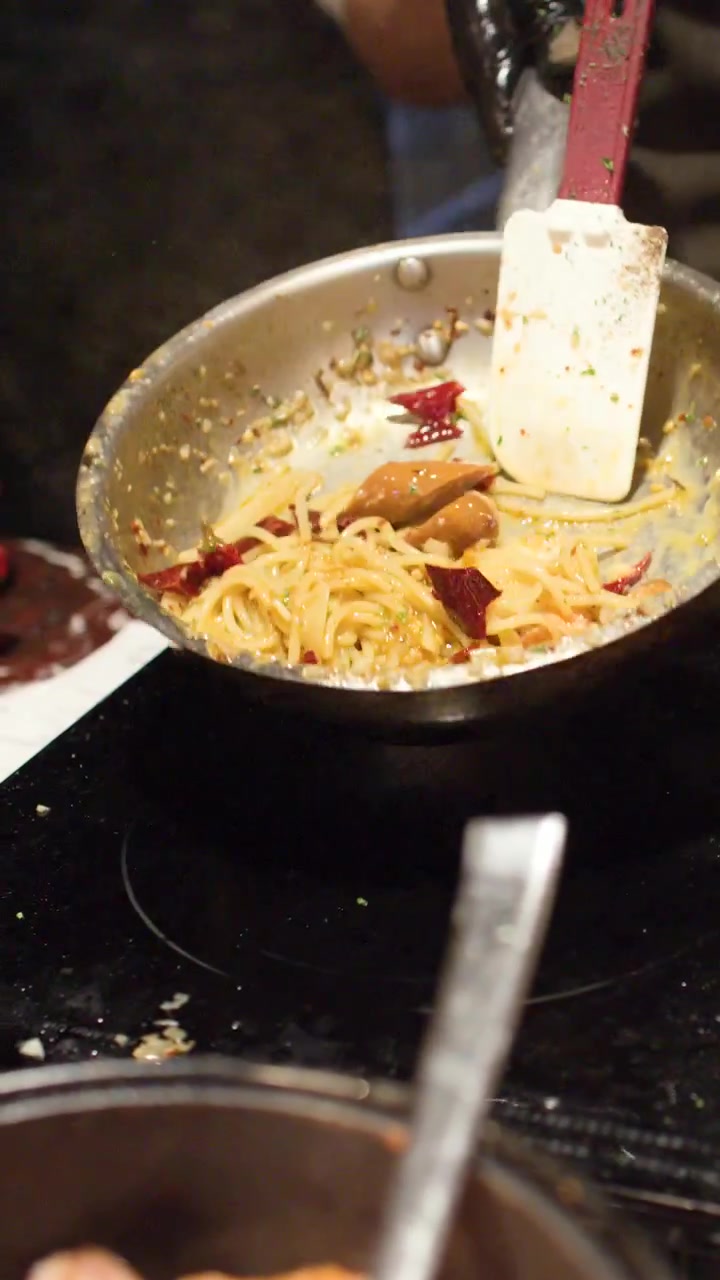 Uno chef salta gli spaghetti in una padella di acciaio inossidabile usando una spatola bianca.