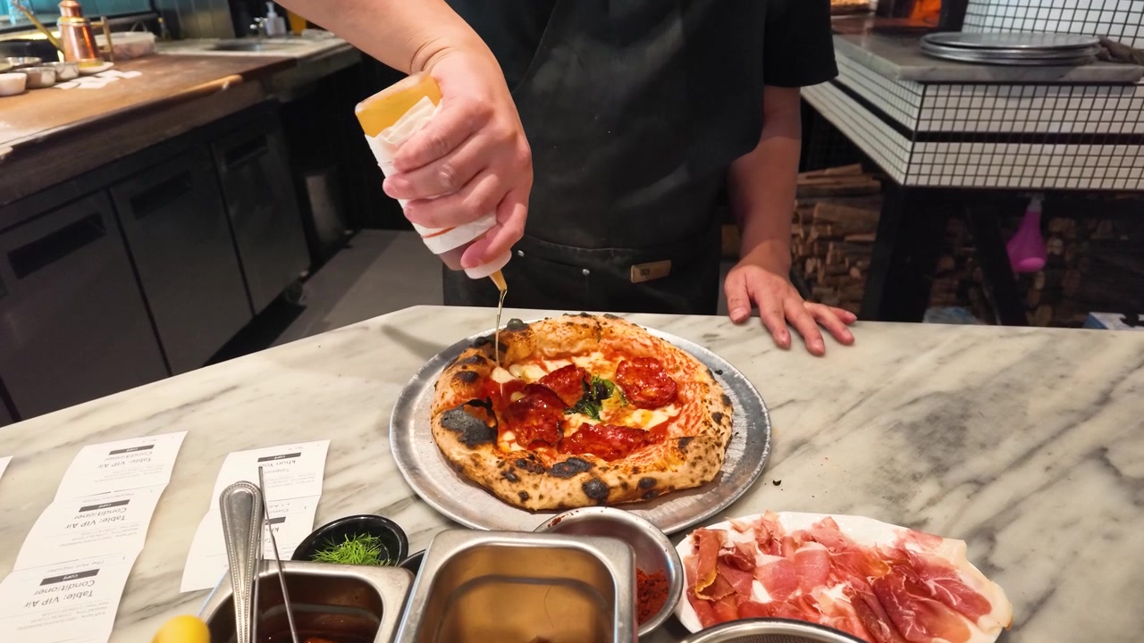 Chef asiatico con grembiule nero aggiunge salsa a una pizza al salamino piccante su un piano di lavoro in marmo in cucina