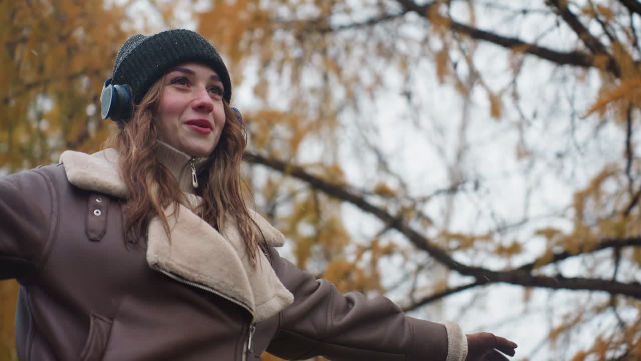 Close view of smiling young girl wearing black knit cap, brown shearling jacket with arms outstretched, walking joyfully outdoors on cool autumn day, embracing nature and fresh air