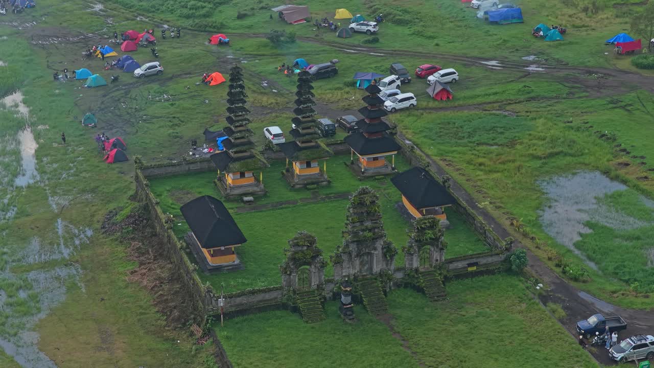Geleneksel Bali Hindu tapınağı Pura Ulun Danu Tamblingan'ı gösteren yüksek açılı görünüm, arabalar ve çadırlarla dolu canlı bir kamp alanı ile çevrili.