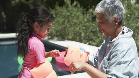 Una-Chica-Birracial-Con-Un-Traje-De-Baño-Rosa-Juega-Con-Alas-De-Agua,-Sonriendo-A-Su-Abuela