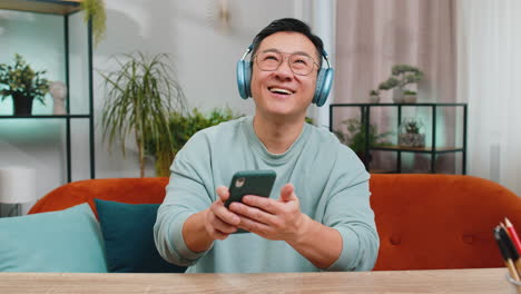 happy relaxed man asian in headphones dancing listening favorite rock n roll music on home couch