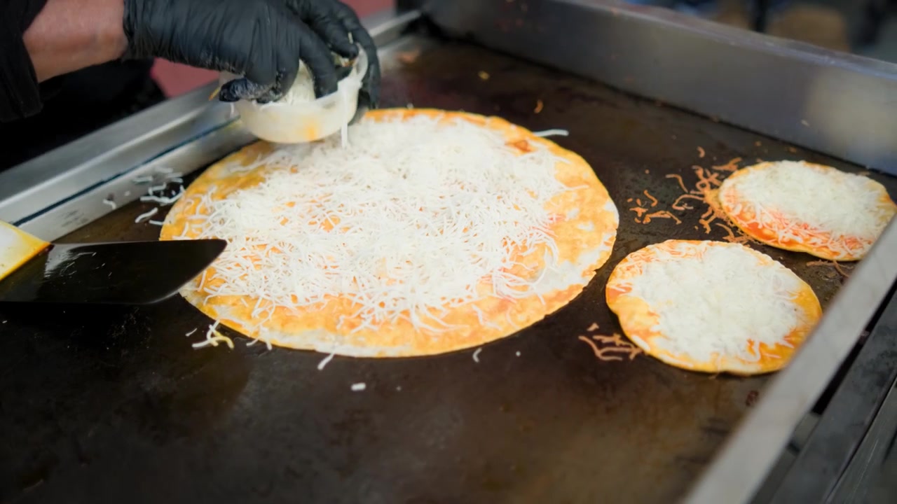 Rekaman dapur komersial menunjukkan penambahan keju pada pizza birria bersama beberapa quesabirria selama persiapan pizza birria gurih di atas panggangan datar