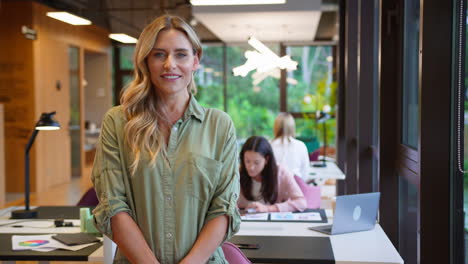 Portrait-Of-Smiling-Businesswoman-Standing-By-Desk-In-Modern-Open-Plan-Office