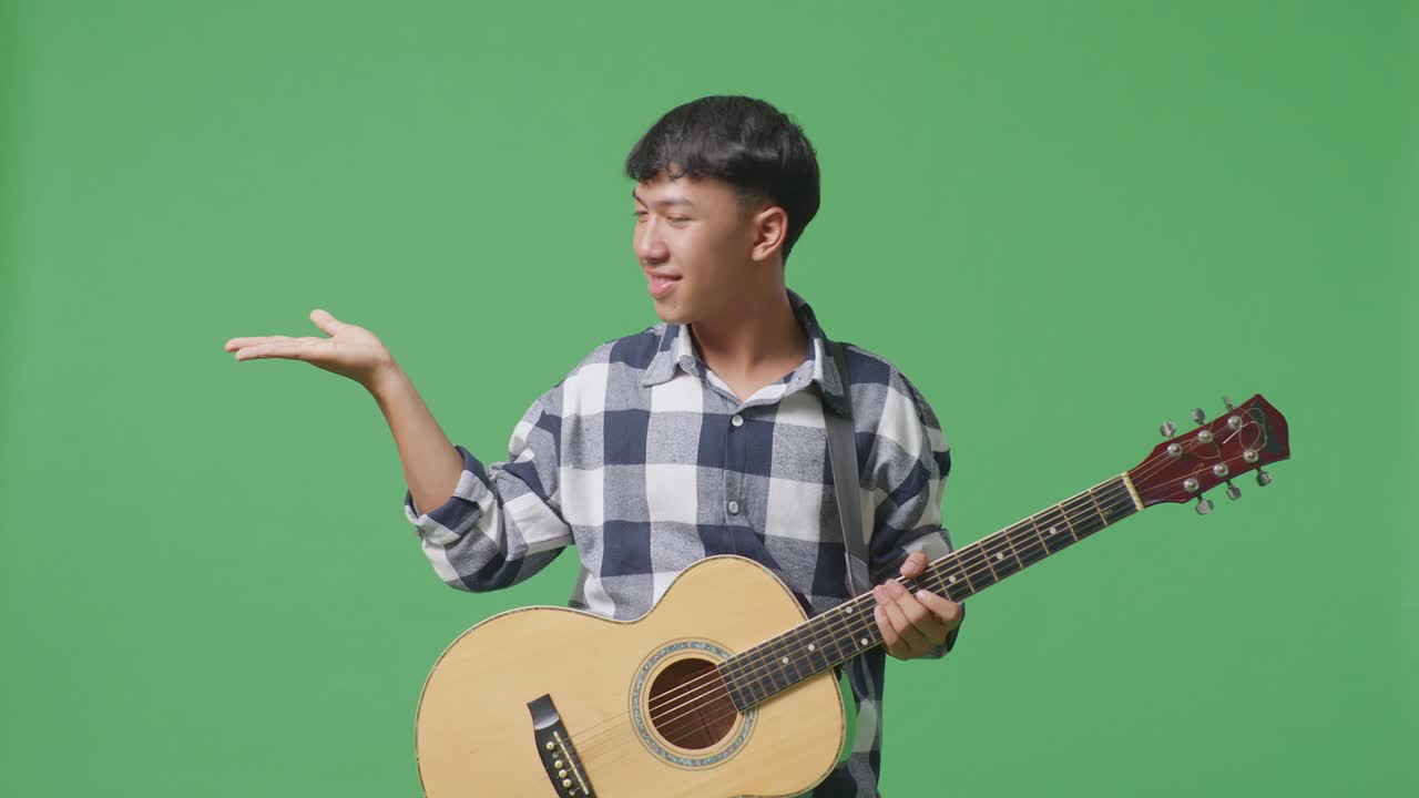 기타를 들고 있는 아시아 십대가 미소 짓고 녹색 화면 배경에서 을 가리키고 있다