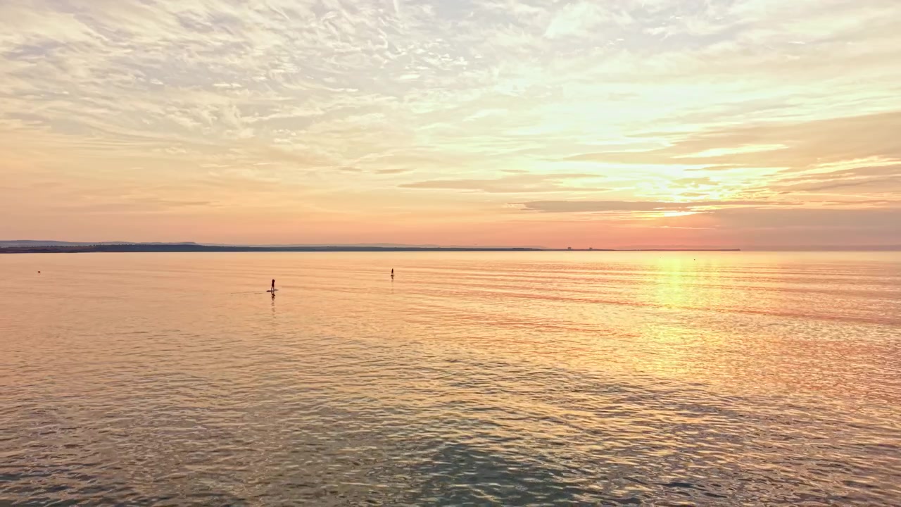 Sunset view over calm water with silhouettes of two people stand-up paddle boarding during twilight hours