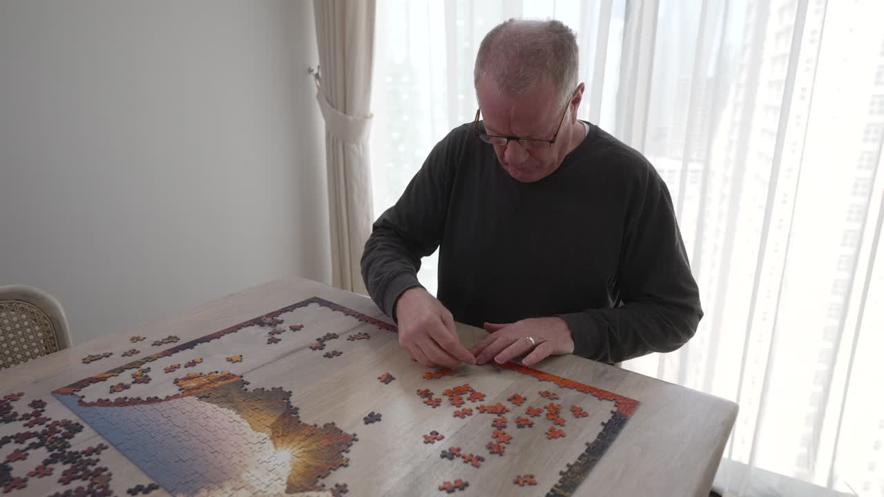 Senior man with focused determination puts a large puzzle together in his lonely apartment - pullback reveal