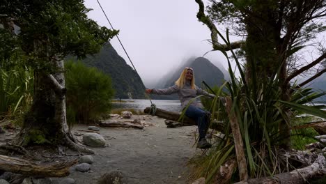 Cheerful-blonde-woman-on-a-swing-in-Milford-Sound,-New-Zealand