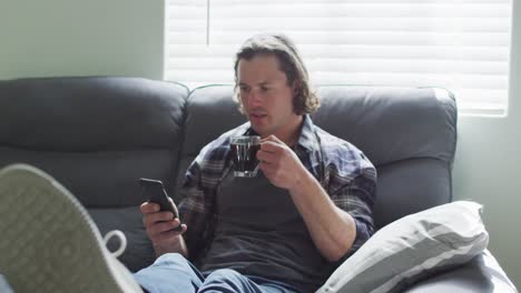 hombre caucásico feliz sentado en el sofá en la sala de estar usando un teléfono inteligente y bebiendo café
