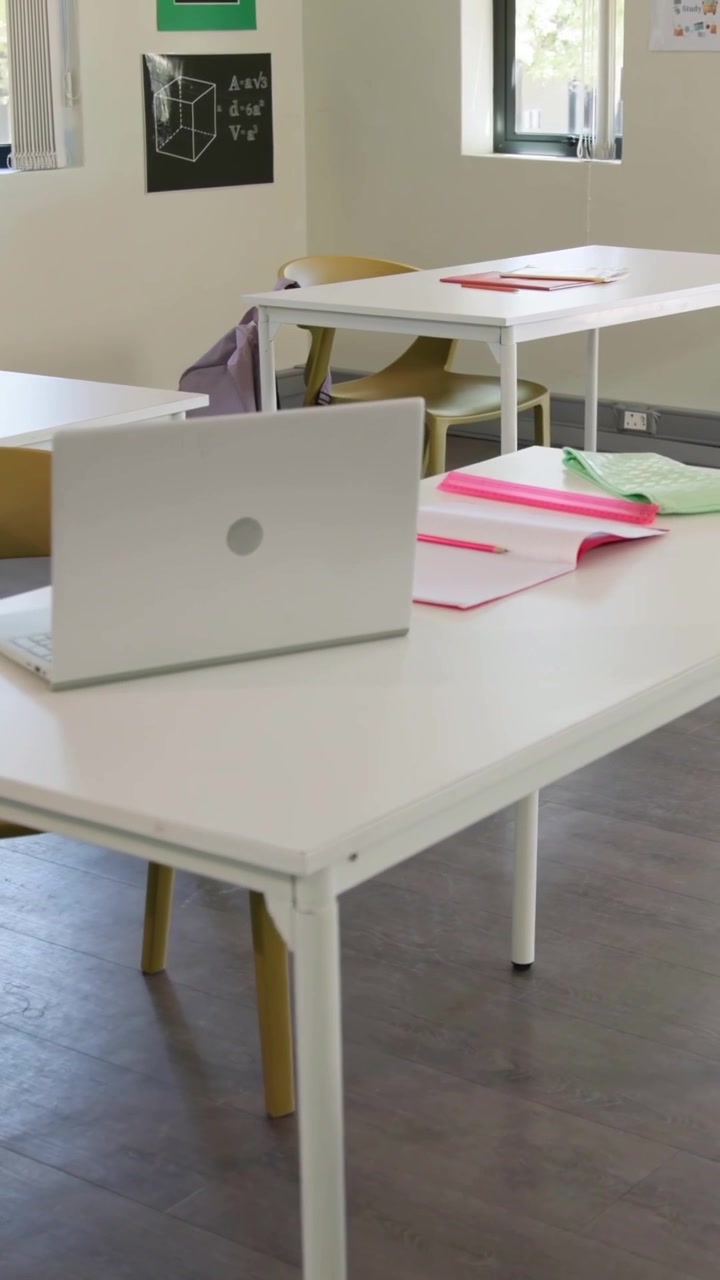 Vertical video: Panning camera revealing silver laptop lid on table in classroom with notebooks