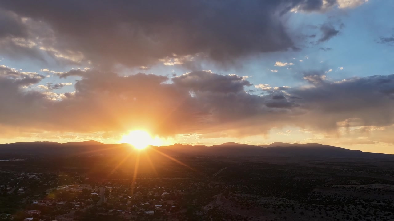 4K UHD drone timelapse capturing a glowing sunset over a suburban town. The brilliant sun dips behind a silhouetted mountain range, casting warm light beneath broken, heavy evening clouds.