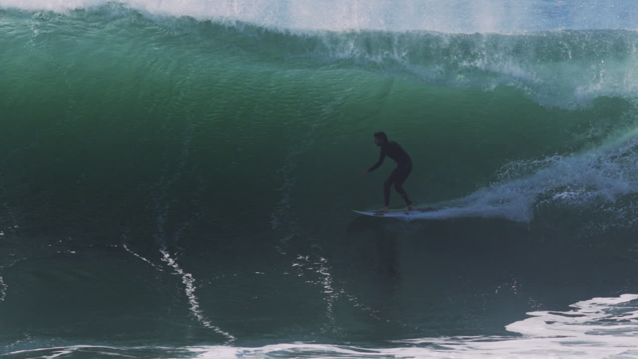 Surfer drops into massive green barrel as wave curls and sunlight hits translucent water