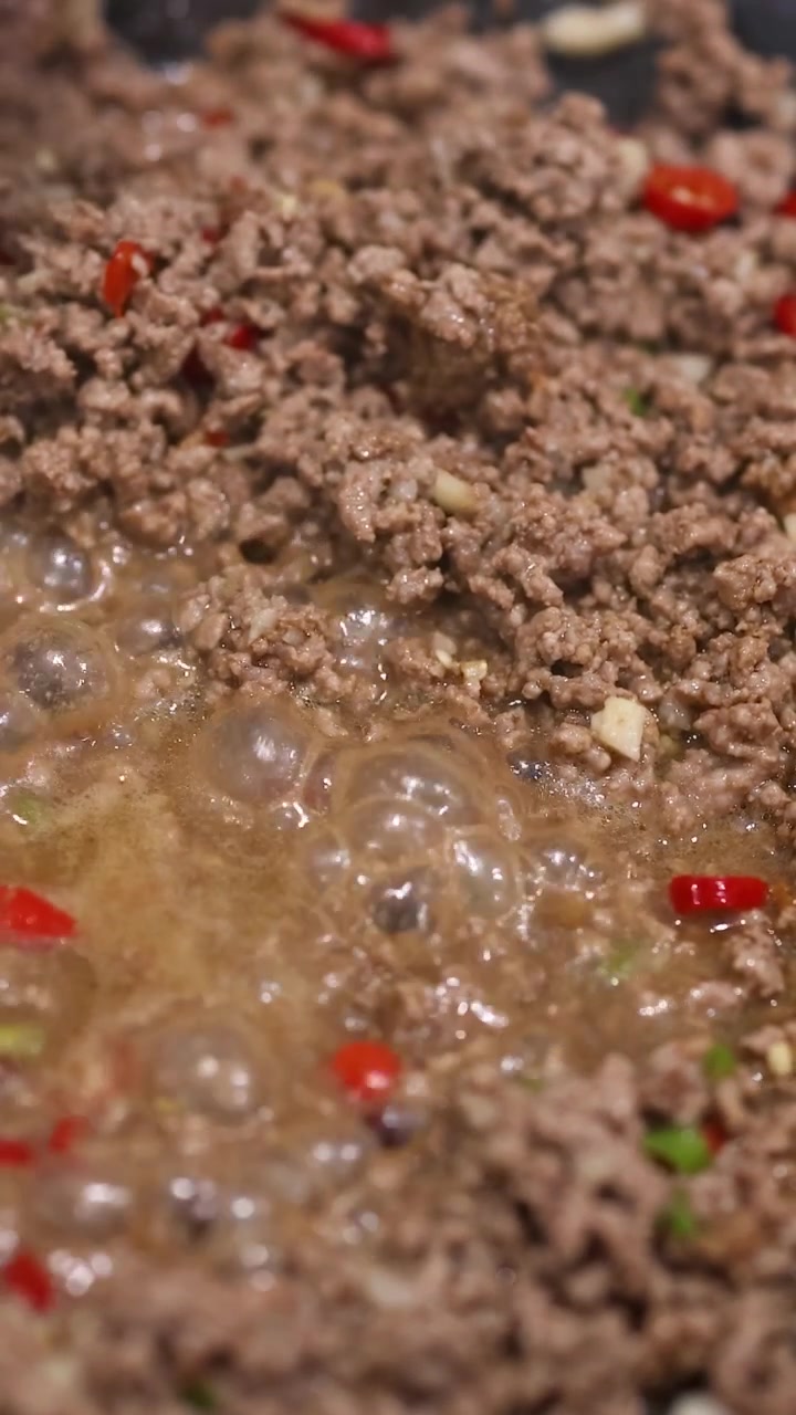 Daging sapi giling dimasak dengan paprika merah dan rempah-rempah dalam saus yang mendidih. Tampilan jarak dekat menangkap proses memasak yang semarak.