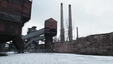 old, abandoned industrial complex covered in snow, with rusted structures, buildings and tall chimneys standing against an overcast sky