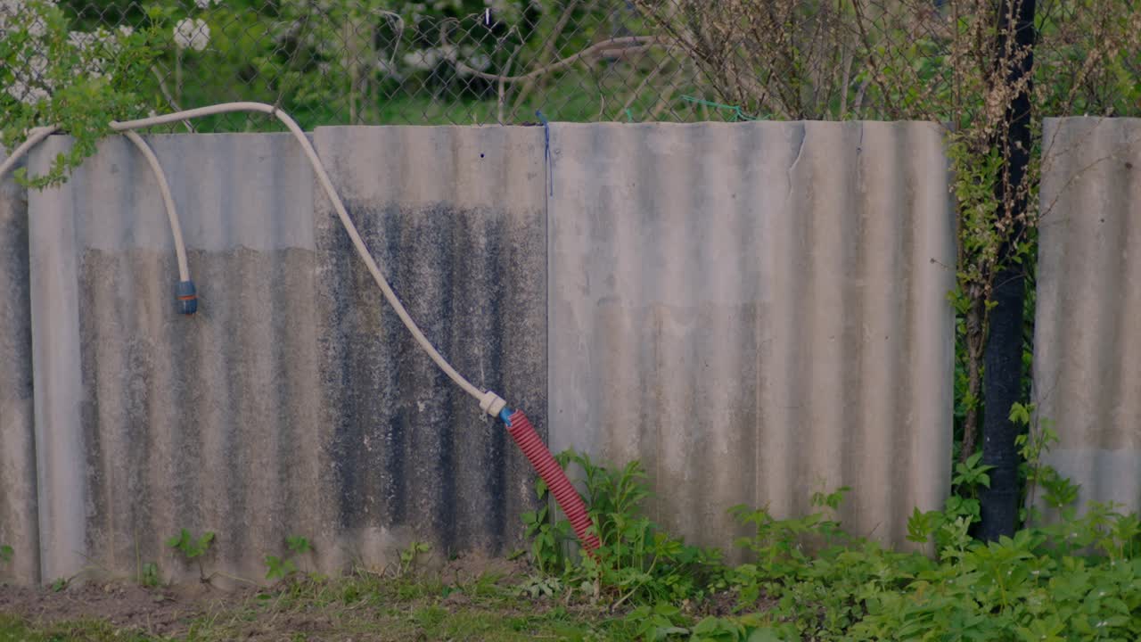 Old, corrugated metal fence with a hose draped over it, set in a rural, overgrown environment. The scene reflects an aged, rustic aesthetic, featuring weathered metal, greenery, and a makeshift setup.