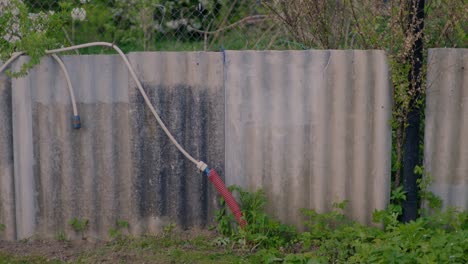 old, corrugated metal fence with a hose draped over it, set in a rural, overgrown environment. the scene reflects an aged, rustic aesthetic, featuring weathered metal, greenery, and a makeshift setup.