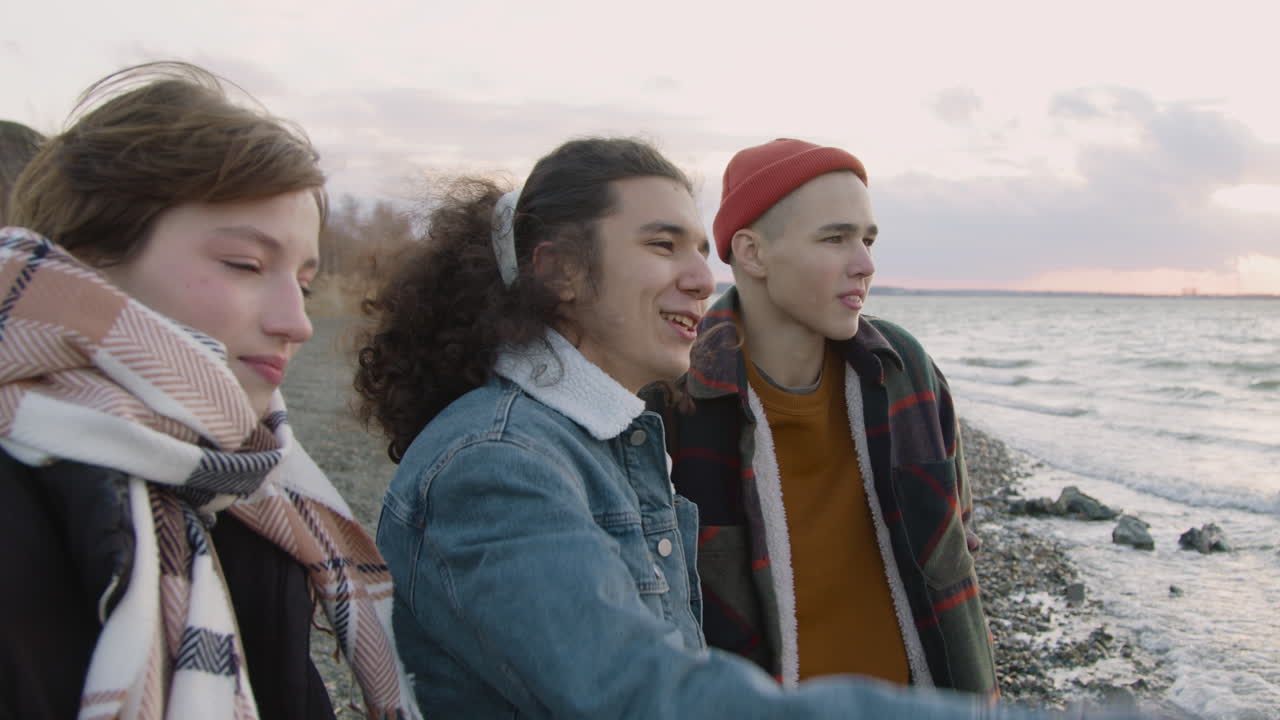 tres amigos adolescentes vestidos de invierno a la orilla del mar hablando, sonriendo y mirando el océano en un día ventoso