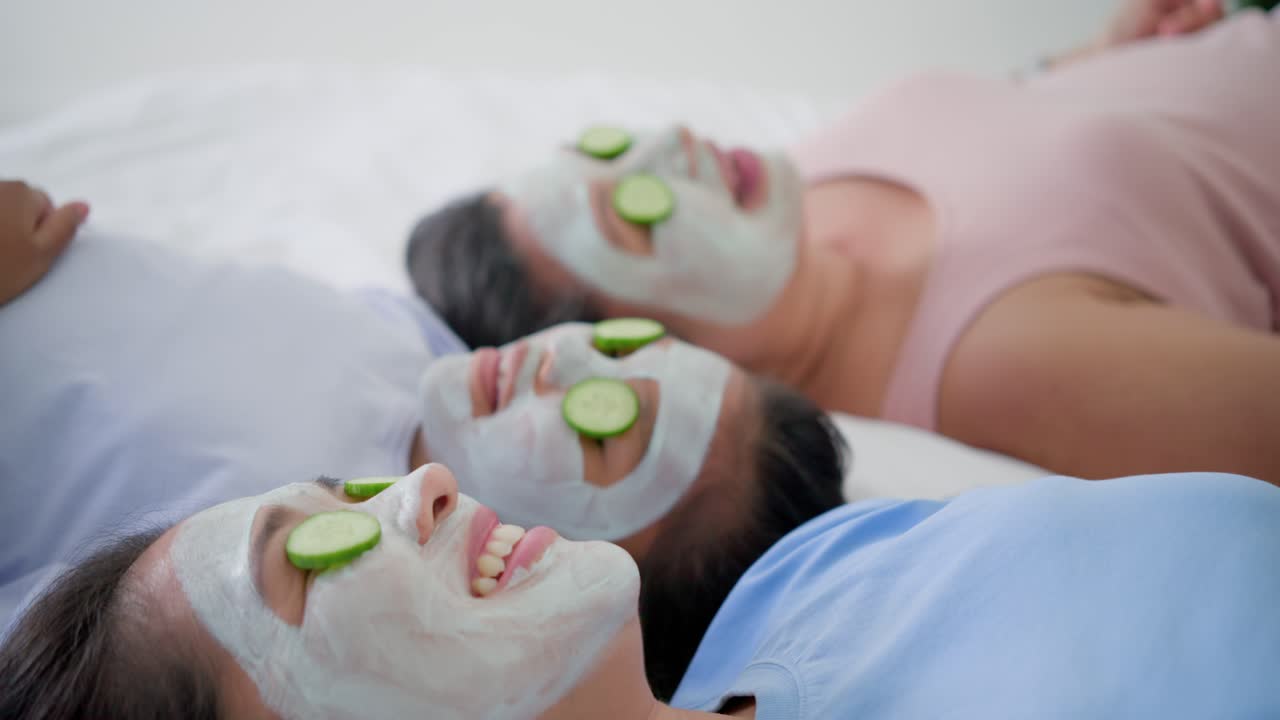 Madre e due figlie di diverse etnie sdraiate sul letto con la maschera, mani da destra posizionano cetrioli per il riposo.