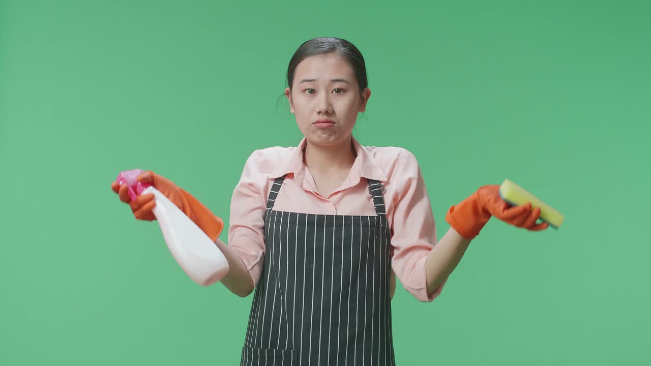 초록색 스크린 스튜디오에 서 있는 스프레이와 부 스폰지를 보여주는 치마를 입은 혼란스러운 아시아 여성 집주인