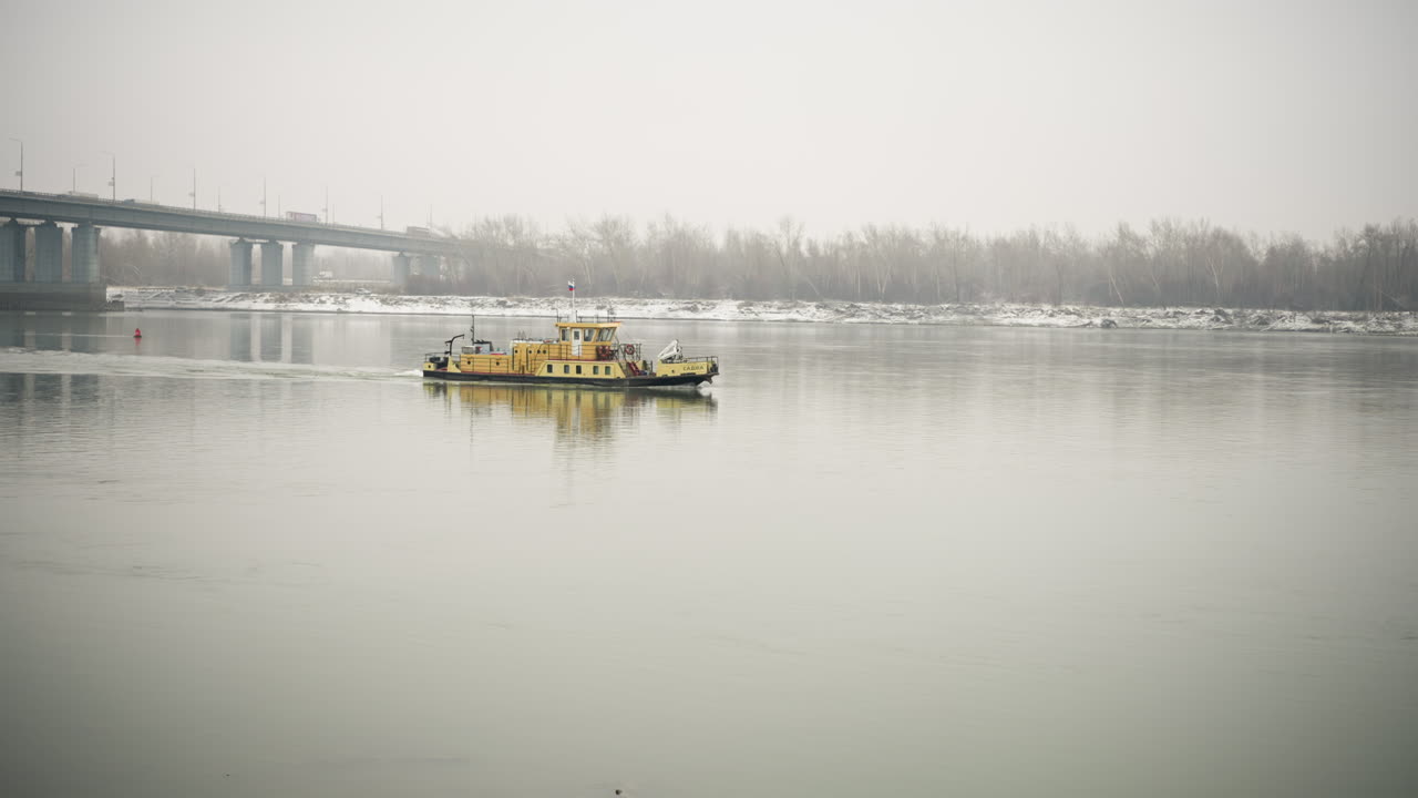 노란색 배는 안개가 가득한 강에서 조용히 항해하고 나무의 반사와 겨울의 분위기에서 먼 다리를 둘러싸고, 평온한 물과 해변에 가운 눈이 내리고, 구름이 가득한 하늘 아래