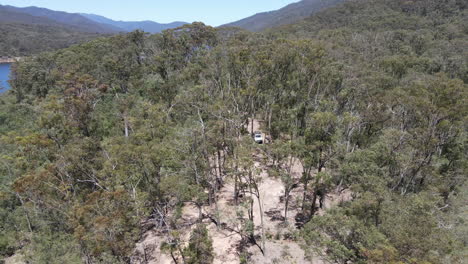 Aerial-drone-shot-of-4WD-car-Moving-away-to-show-tall-trees,-green-bushland,-and-blue-water-near-Lake-Eildon,-Victoria-Australia