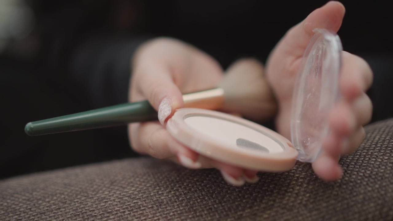 Close view of manicured hands holding compact and makeup brush while softly applying powder, showing careful fingertip movement, beauty routine detail, and clean studio lighting, with makeup, brush