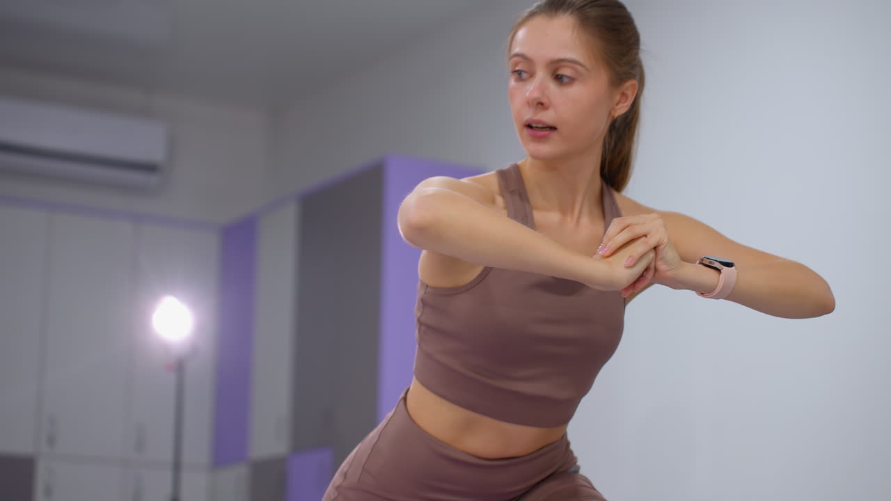 mujer con ropa deportiva marrón con las manos unidas se centra en el ejercicio de cuclillas, se baja en un movimiento controlado, manteniendo el equilibrio y la postura con equipos de gimnasia en el fondo