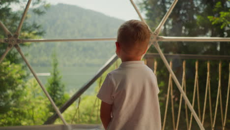 child looks around out of glass dome at glamping. little boy spellbound by landscape with lake near panoramic window. kid enjoys summer forest scene