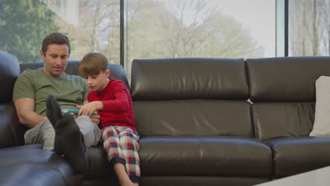 father with son wearing pyjamas sitting on sofa at home playing computer game on handheld device - shot in slow motion