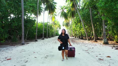 mujer asiática emocionada que llega a la isla paradisíaca con la maleta con ruedas en ropa informal