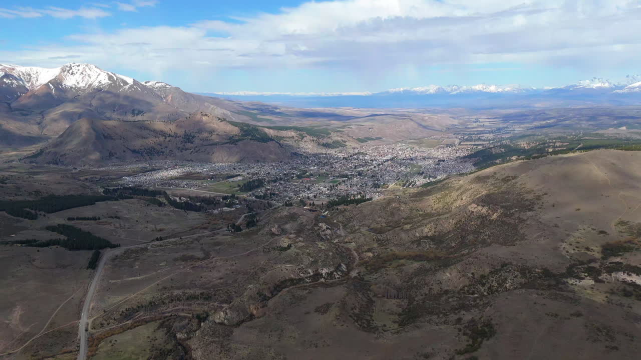 Daisy cutter drone shot over Esquel town in Argentina south. Dry arid hills make good contrast with snow in mountain range. Detail reveals trees and vegetation on ground. Shot on DJI Air3 at 4K-60fps.