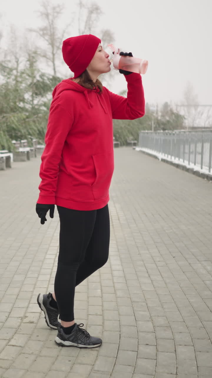 Lady in pose opening water bottle and sipping water from pink bottle outdoors in serene winter park setting with snow-covered ground, iron railing, benches, trees in misty background