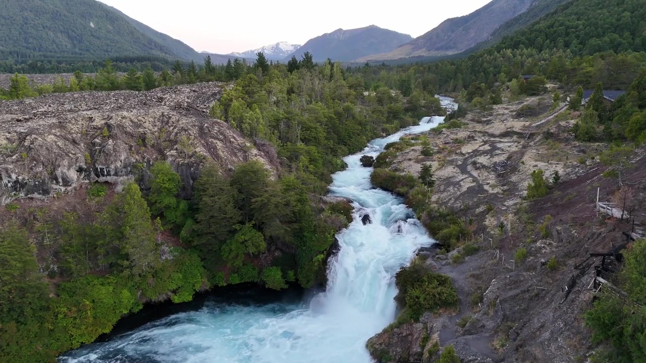 Riprese aeree con drone che si innalzano sopra rapide di acqua bianca e cascate scavate attraverso canyon di roccia lavica scura. Fitta foresta verde e sfondo di vulcano innevato sotto un cielo leggermente coperto.