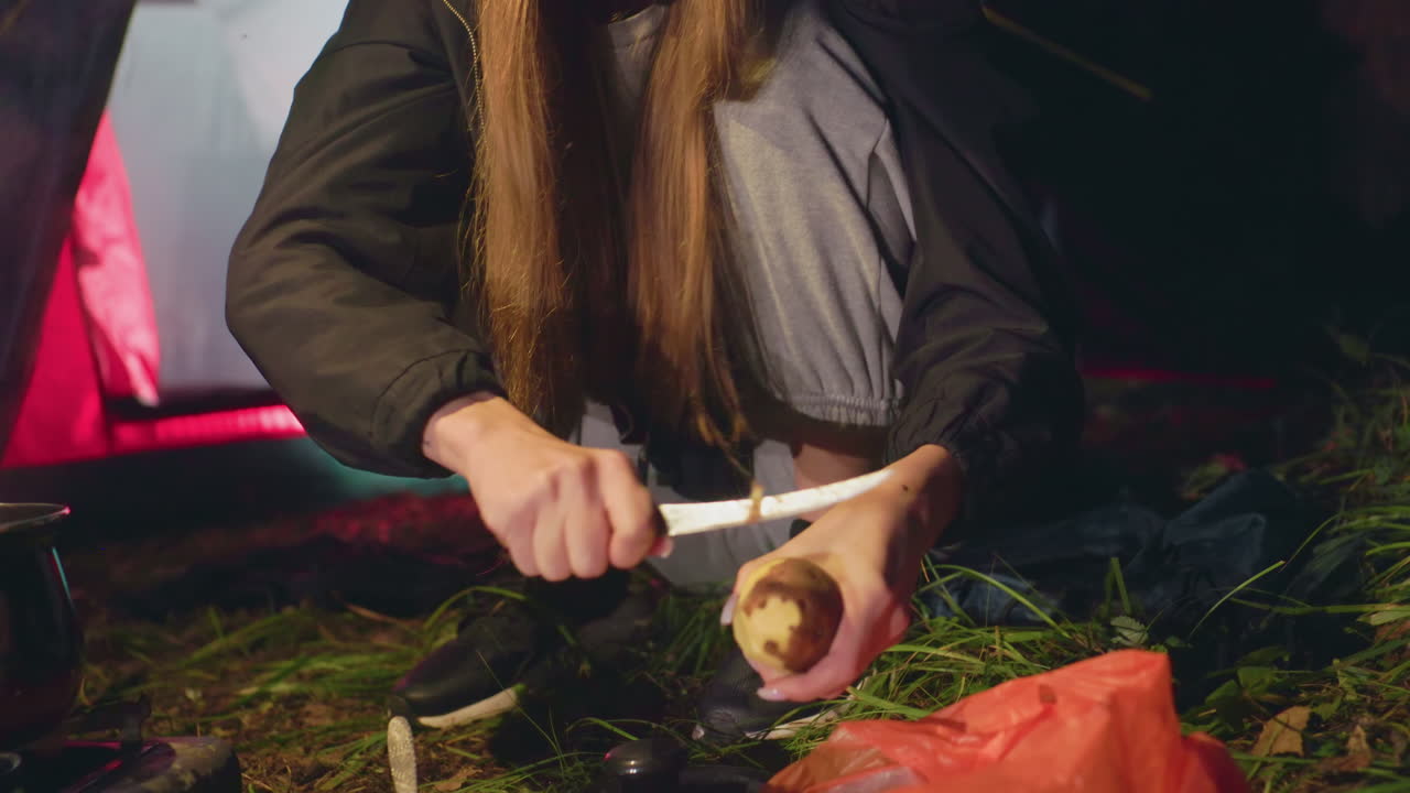 Close up of young lady peeling potatoes beside tent during night camping in forest, dressed in warm clothes with beanie, focused on food preparation while sitting on grass in calm outdoor environment