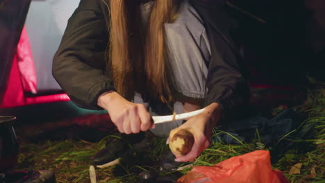 close up of young lady peeling potatoes beside tent during night camping in forest, dressed in warm clothes with beanie, focused on food preparation while sitting on grass in calm outdoor environment