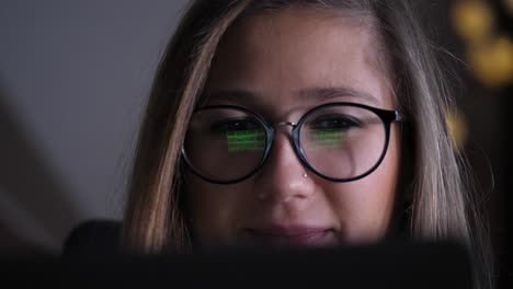 una cara de mujer joven que se muestra detrás de una pantalla, está desplazando imágenes en el navegador, la pantalla se refleja en sus gafas, sonríe que está enfocada