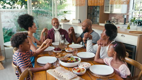 multi-generation family joining hands and saying prayer before meal at home