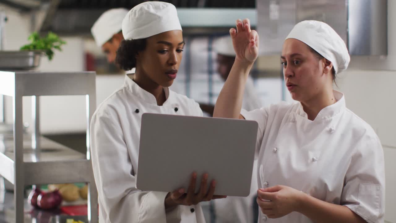 dos cocineras diversas hablando y usando una computadora portátil en la cocina del restaurante