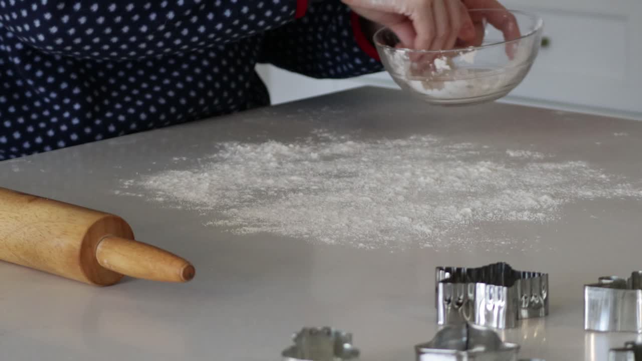 A close up of flour being sprinkled on a table next to a rolling pin and cookie cutters in preparation to bake cookies