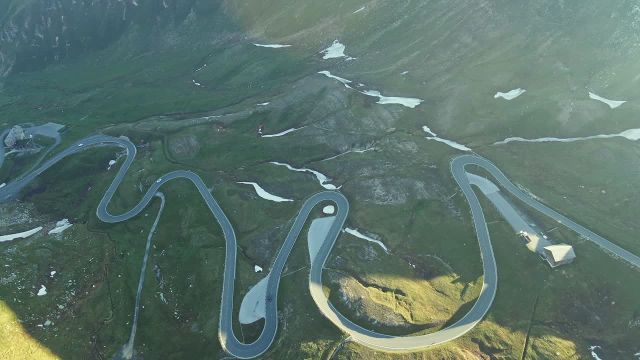 Aerial view of a winding mountain road with sharp switchbacks cutting through a rugged alpine landscape. Patches of lingering snow and golden sunlight highlight the dramatic terrain.
