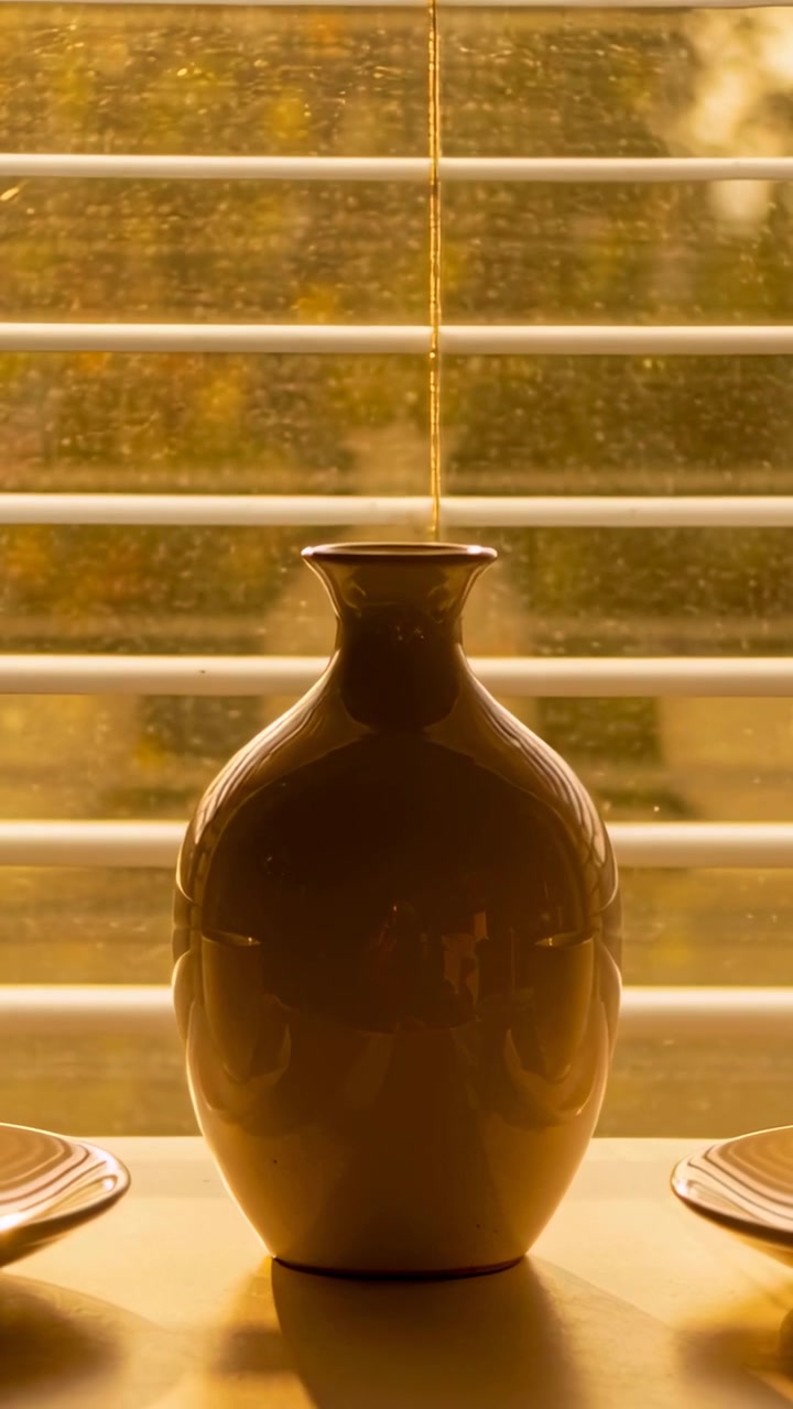 Vidéo verticale : Lumière du soleil changeante, un vase reflétant une lumière chaude sur le rebord de fenêtre, montrant des stores, un cordon et des tasses.