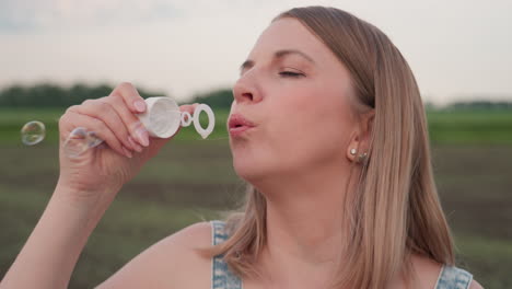 close up of rosy cheeked young woman blowing soap bubbles with wand near lips under pastel sky over lush field with iridescent bubbles glinting in soft evening light