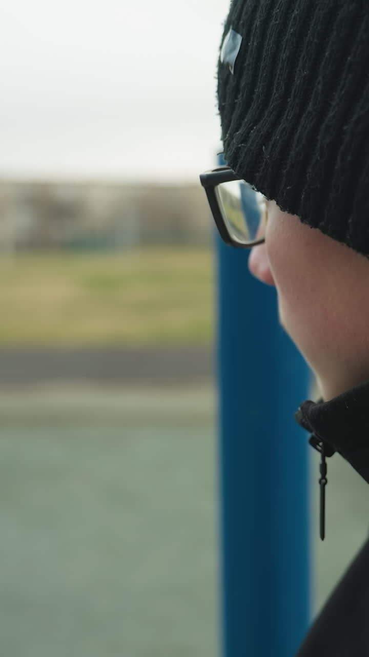 Close-up of a young boy standing near a blue pole, looking thoughtful as he gazes away toward the field, with a blurred view of someone standing by the track and people passing by