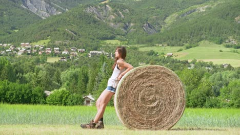 A-beautiful,-strong-brunette-woman-taking-a-break-from-her-work-and-resting-against-a-large-bale-of-hay-on-a-farm-in-France