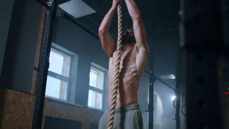 a man in the gym climbs up the rope to the top. perseverance and strength. a sweaty man without a t-shirt is training.